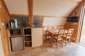 Uma cozinha compacta com pia, micro-ondas e pequeno frigorífico. Há uma mesa de jantar de madeira com cadeiras e uma TV fixada na parede.