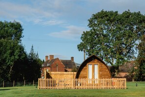 A wooden cabin with a curved roof, surrounded by a wooden fence, set in a grassy area with trees and a brick house in the background.