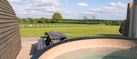 A hot tub with a wooden deck and a picnic table, overlooking a fenced yard with trees and a clear sky.