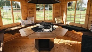 Una cabaña de madera con una mesa central, una parrilla, ventanas amplias y una zona de estar acogedora.