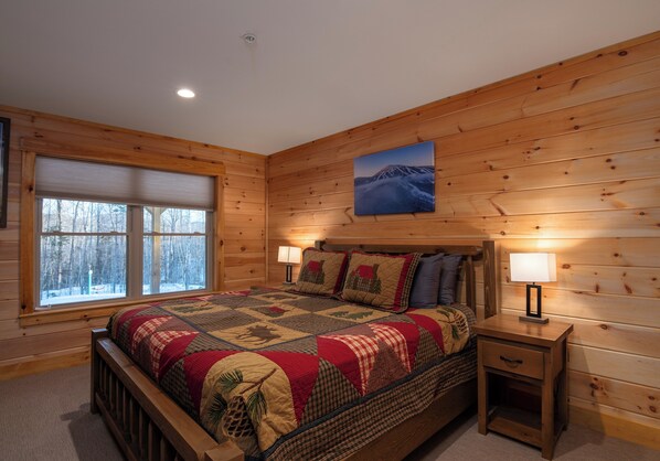 Une chambre confortable avec un mur en bois brut, un grand lit recouvert d’une courtepointe, deux lampes de chevet et une fenêtre donnant sur la forêt.
