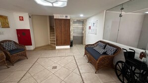 A room with two wicker sofas, a wheelchair, a red door, an elevator, and a tiled floor. - Apartment in Guarujá-Enseada, leave your car in the garage, just 5 minutes from the beach (Loteamento Joao Batista Juliao Guarujá)
