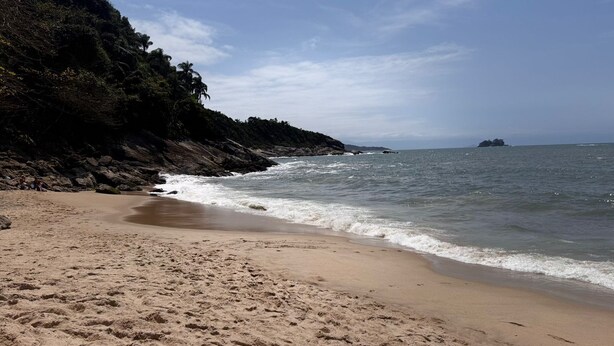 A sandy beach with gentle waves, a rocky cliff, and a distant island. - Apartment in Guarujá-Enseada, leave your car in the garage, just 5 minutes from the beach (Loteamento Joao Batista Juliao Guarujá)