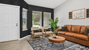 Ein Wohnzimmer mit schwarzer Wand, weißer Tür, zwei beigen Sesseln, einem braunen Ledersofa, einem runden Holztisch und einem gemusterten Teppich.
