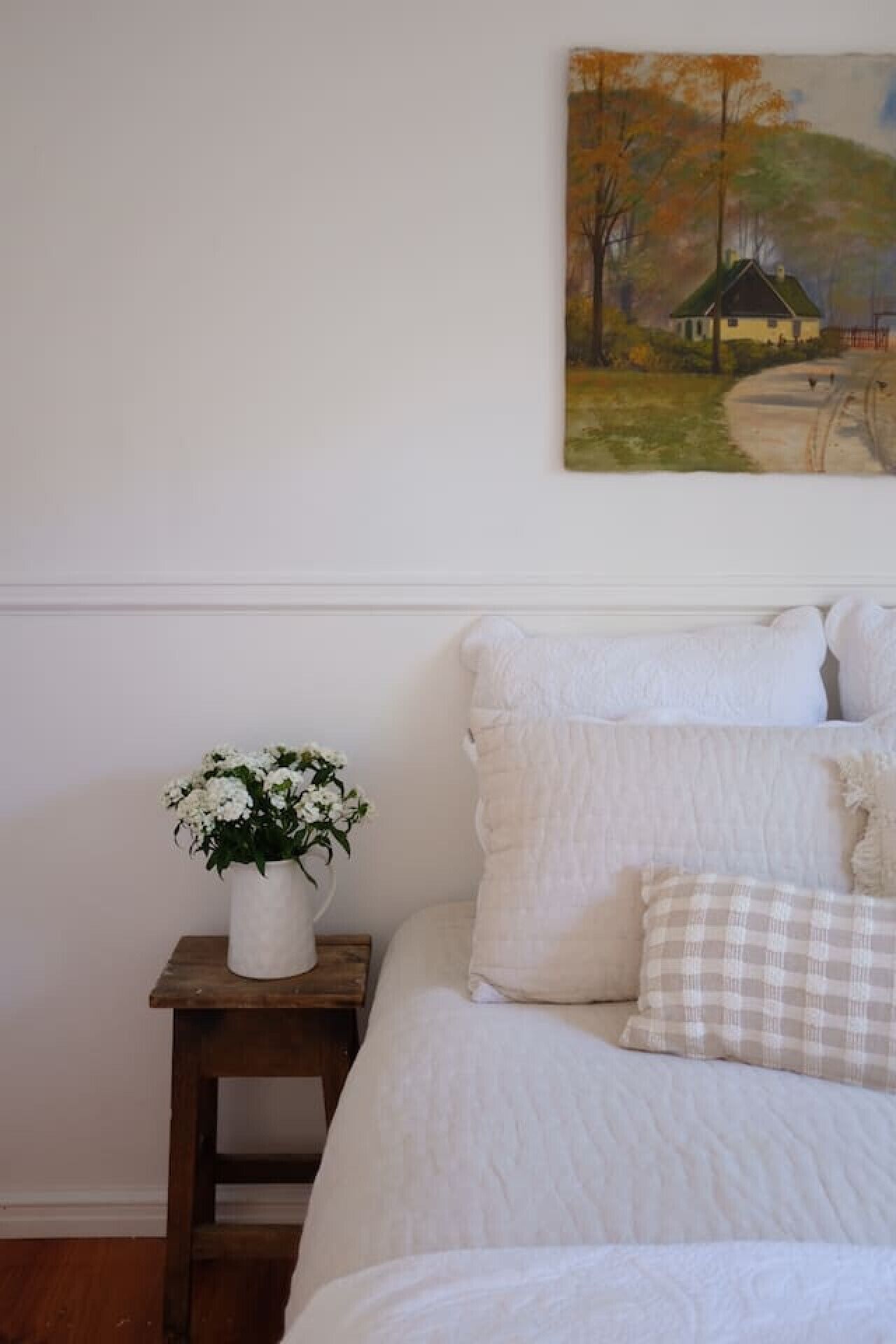 A bedroom with a bed, white bedding, a small wooden nightstand with a white vase of flowers, and a painting of a house on the wall.