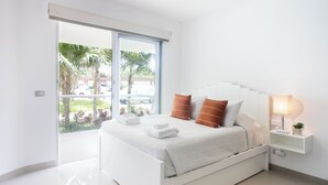 Un dormitorio moderno con una cama blanca, una mesita de noche con lámpara y vistas a palmeras a través de una puerta corredera.