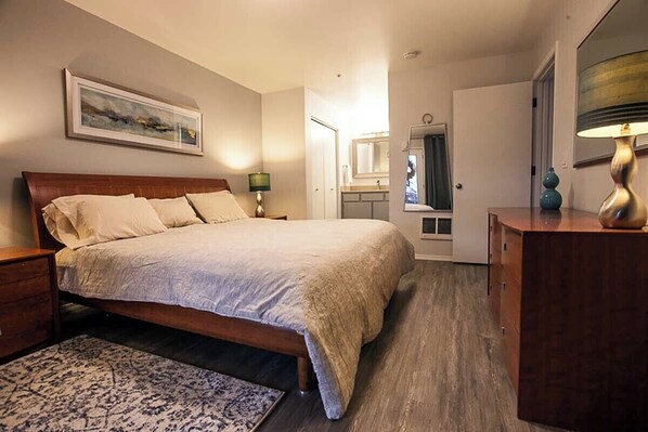 A bedroom with a large bed, a nightstand, a dresser, a mirror, and a door leading to a bathroom.
