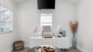 A modern, white room with a flat-screen TV mounted on the wall, a wooden chessboard on a white console, a wicker basket, and a vase with dried plants. - Retreat off Main Street (Safety Harbor)