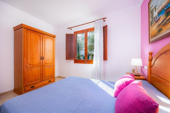 A bedroom with a bed, a wardrobe, a window with curtains, a nightstand with a lamp, and a painting on the wall.