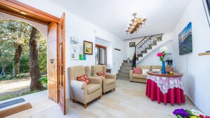 Uma sala ampla com dois poltronas bege, uma porta de madeira que dá para uma área externa, uma escada e uma mesa de jantar com toalha de renda.