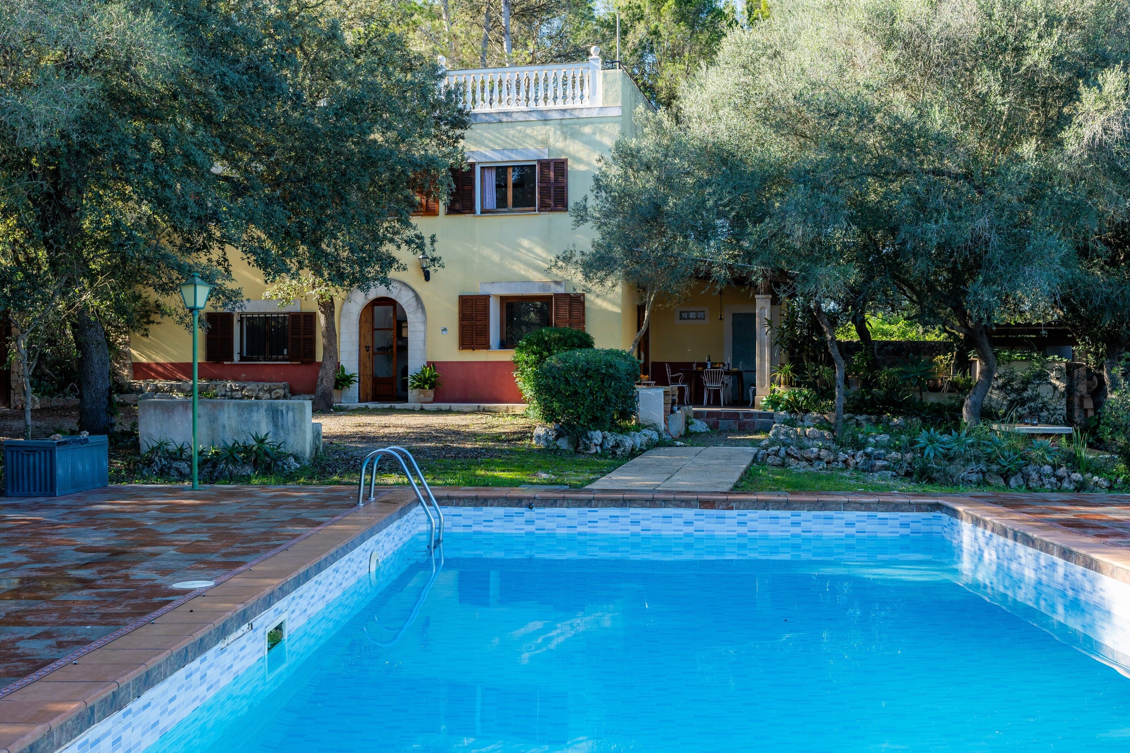 Una piscina con borde de azulejos, rodeada por un jardín con árboles y un edificio con balcón.