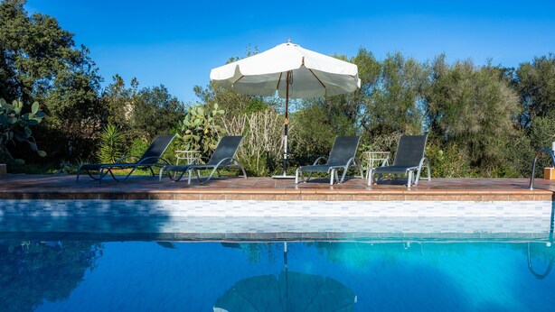 Piscina com água azul cristalina, rodeada por espreguiçadeiras e um guarda-sol branco.