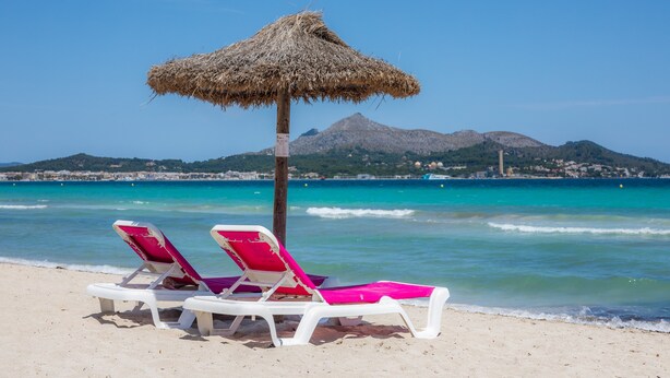 Une plage avec deux chaises longues blanches et un parasol en paille, un ciel bleu dégagé et une montagne en arrière-plan.