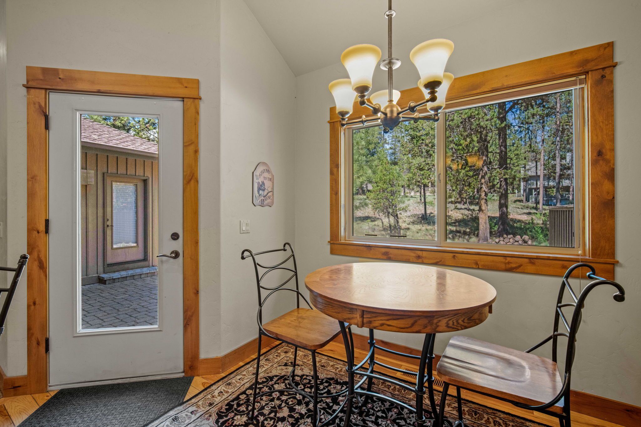 A room with a wooden door, a round table with chairs, a chandelier, and a large window with a view of trees.