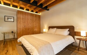 A bedroom with a wooden ceiling, a bed with white bedding, a bedside table with a lamp, and a wooden wardrobe.