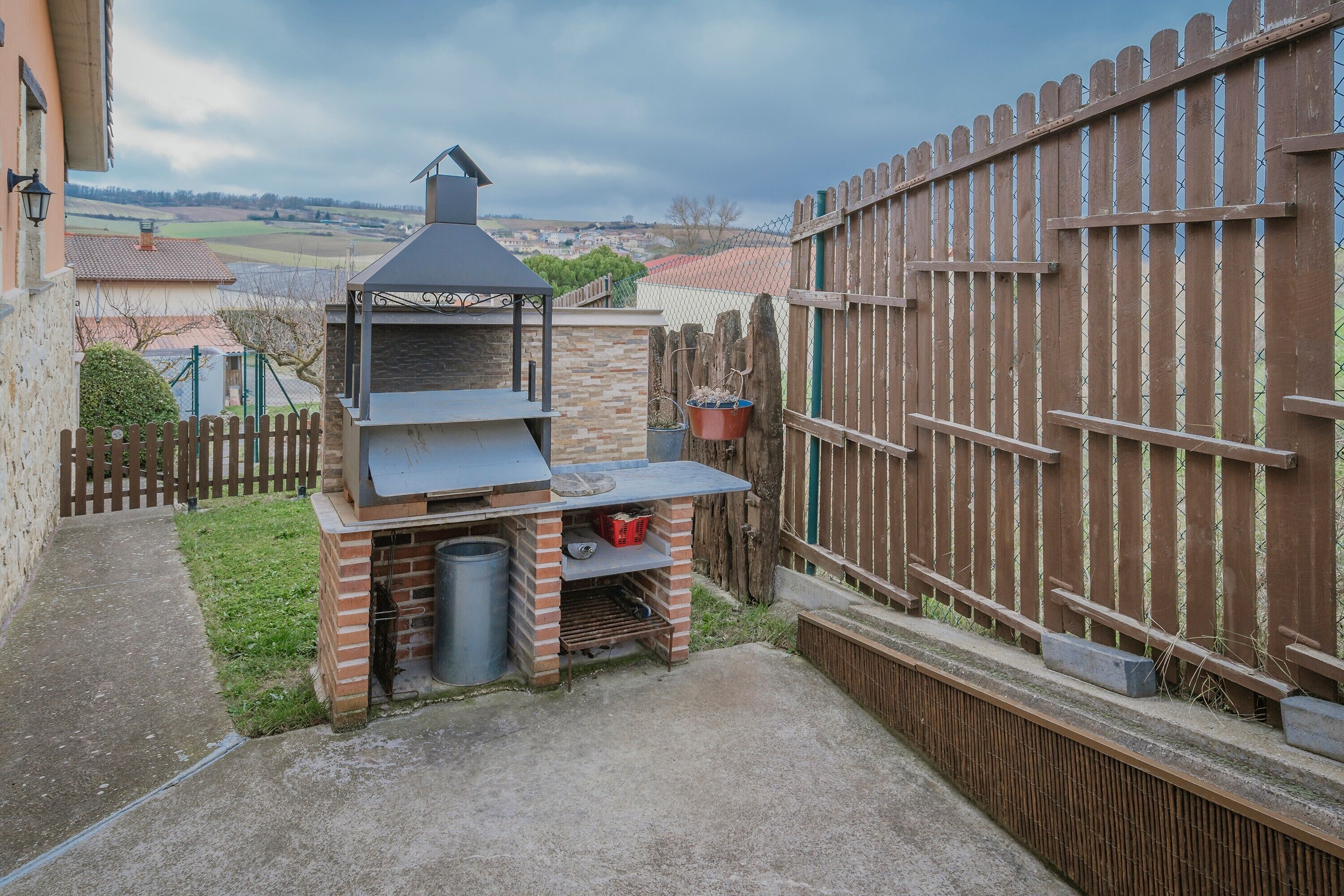 Un patio trasero con una parrilla de ladrillo, una valla de madera y vista al campo.