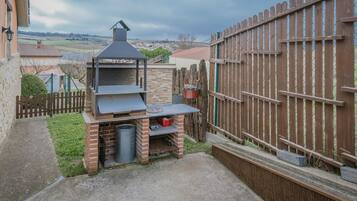 Un patio trasero con una parrilla de ladrillo, una valla de madera y vista al campo.