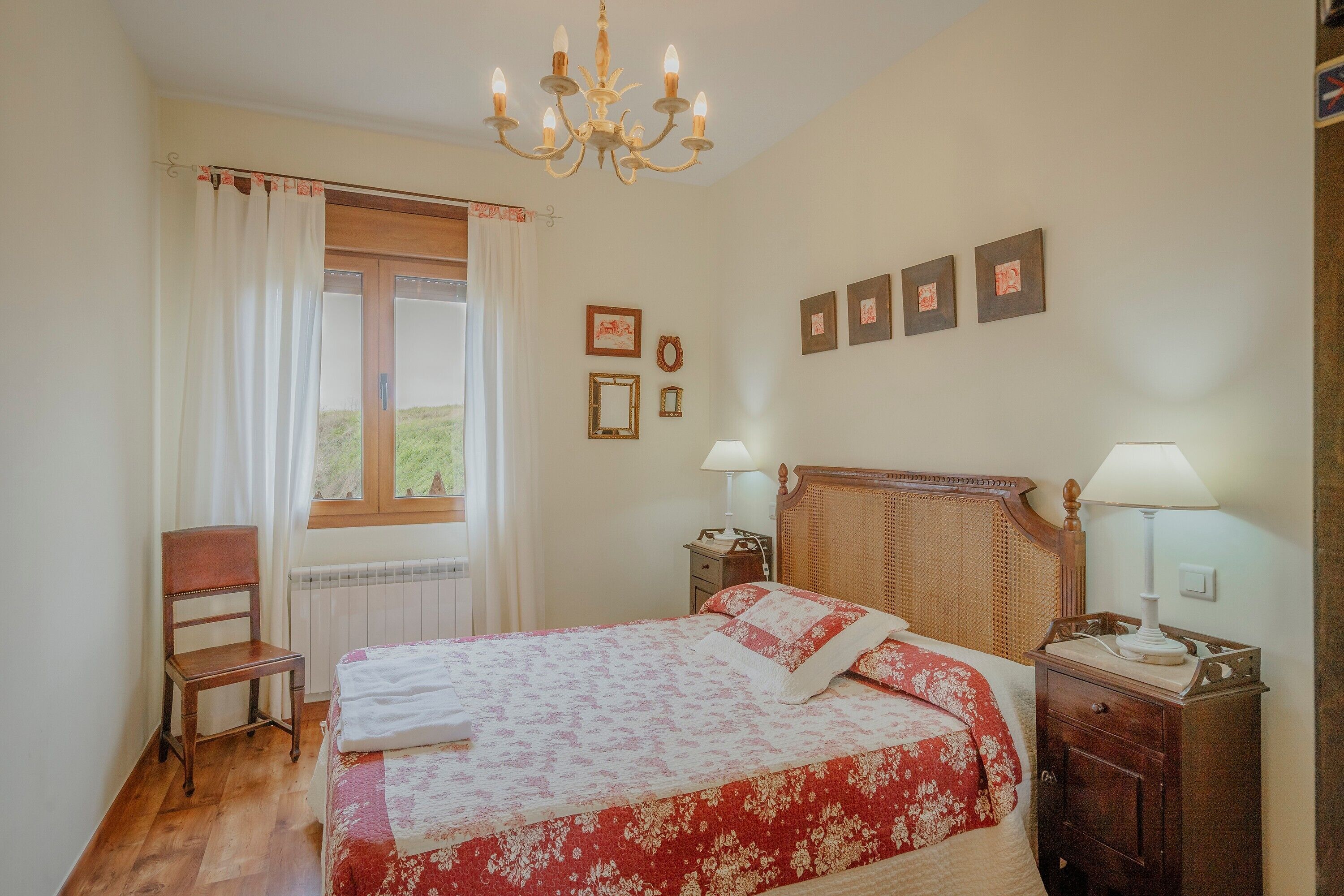 Ein Schlafzimmer mit Bett, Stuhl, Nachttisch, Lampe, einem Fenster mit Vorhängen und Wanddekorationen.