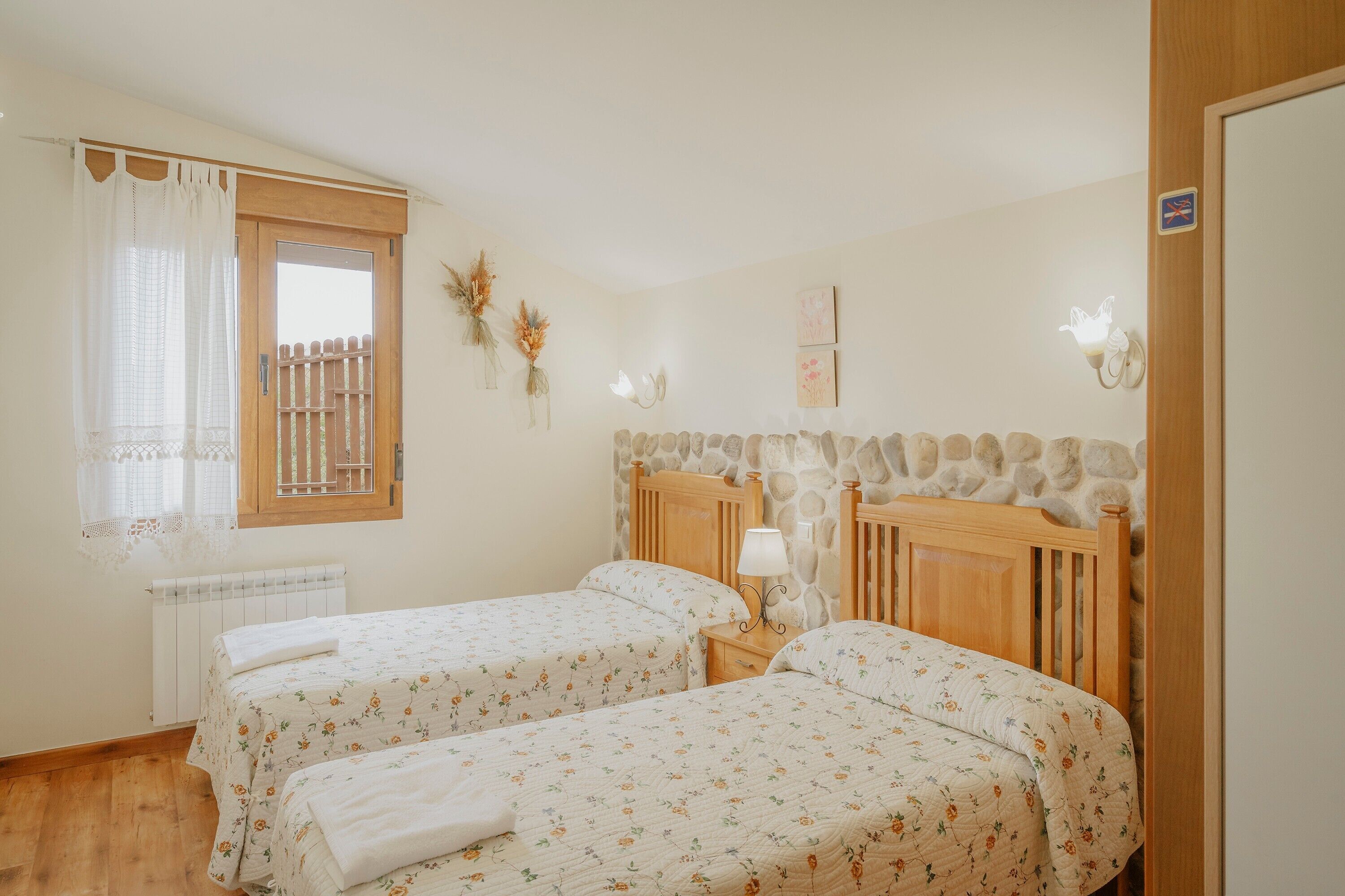 A bedroom with two beds, a window with a curtain, a radiator, a stone wall, and a wooden headboard.