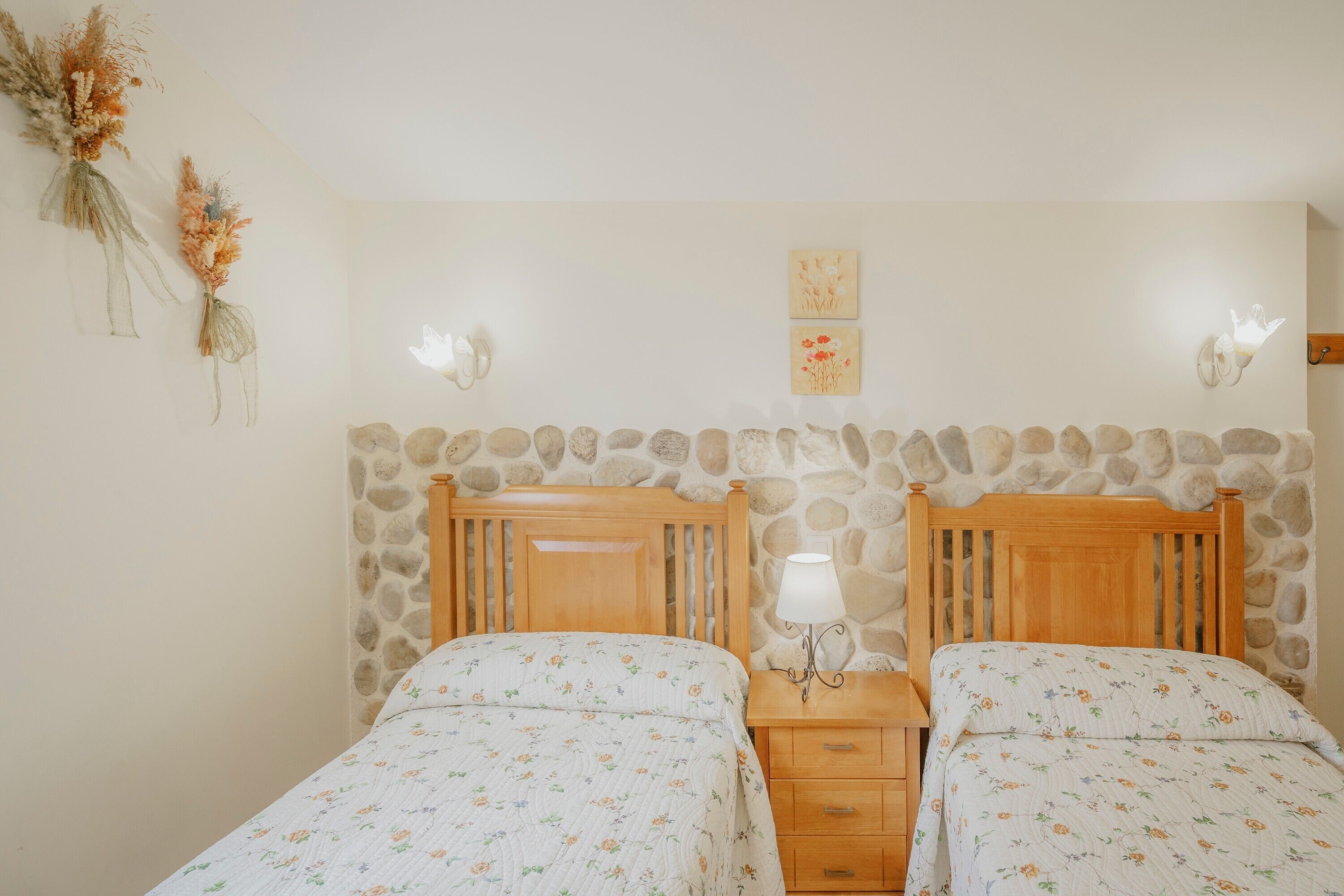 Un dormitorio con dos camas, cabecero de madera, mesita de noche con lámpara y pared de piedra.