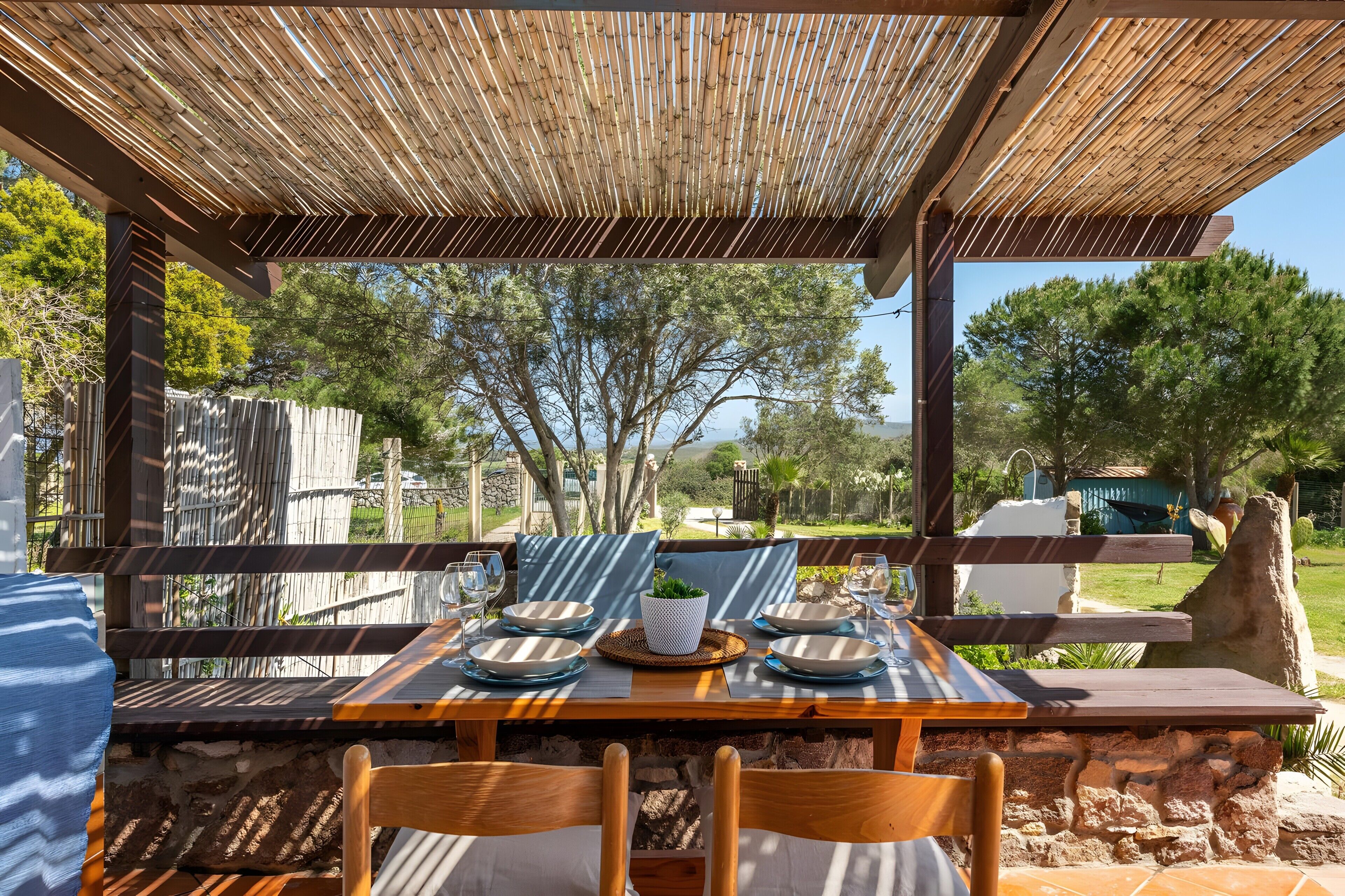Un espace de restauration en plein air agrémenté d’une pergola en bambou, de murs en pierre et offrant une vue sur des arbres et un lac.
