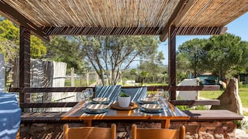 Área de refeições ao ar livre com pérgola de bambu, paredes de pedra e vista para árvores e um lago.
