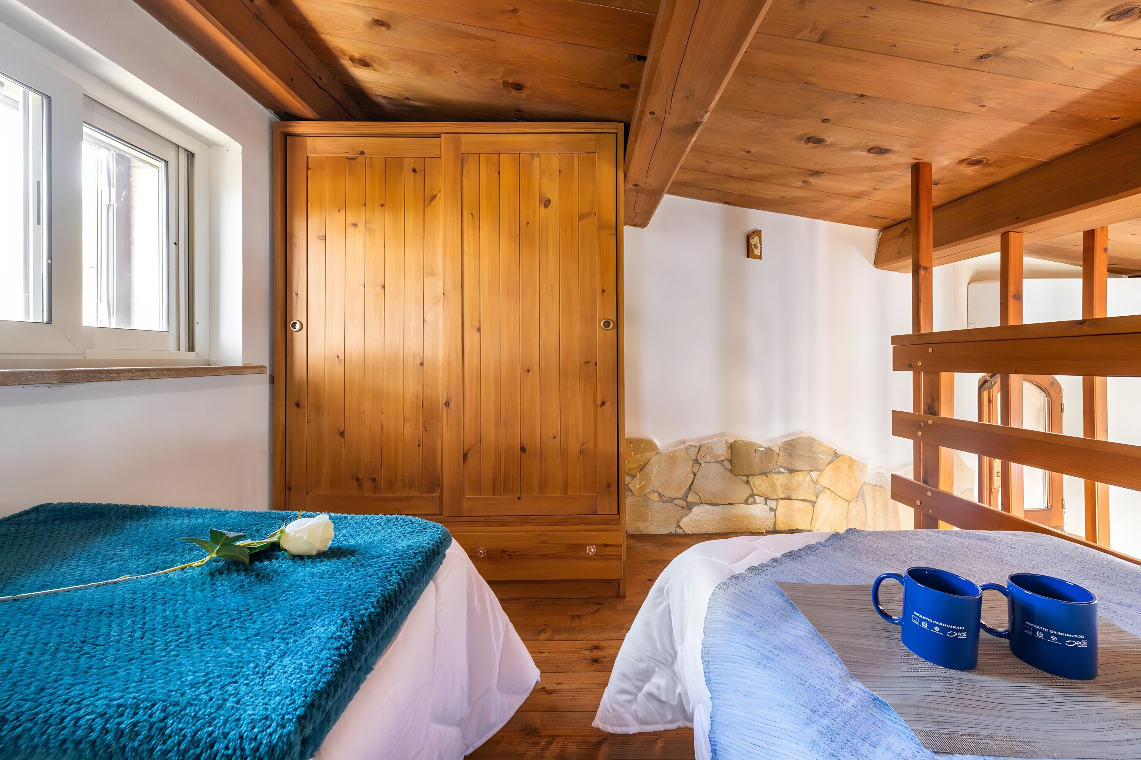 A bedroom with a wooden wardrobe, a bed with a blue blanket, and two blue mugs on a bedside table.