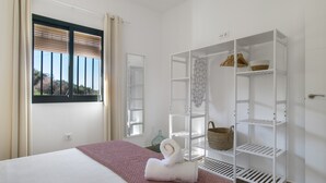 Quarto com cama, janela com persianas, guarda-roupa branco com prateleiras e vista para o verde lá fora.