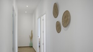 Um corredor com paredes brancas, soalho de madeira e uma porta que dá para outro aposento. Há três decorações de parede trançadas e um vaso verde com juncos.