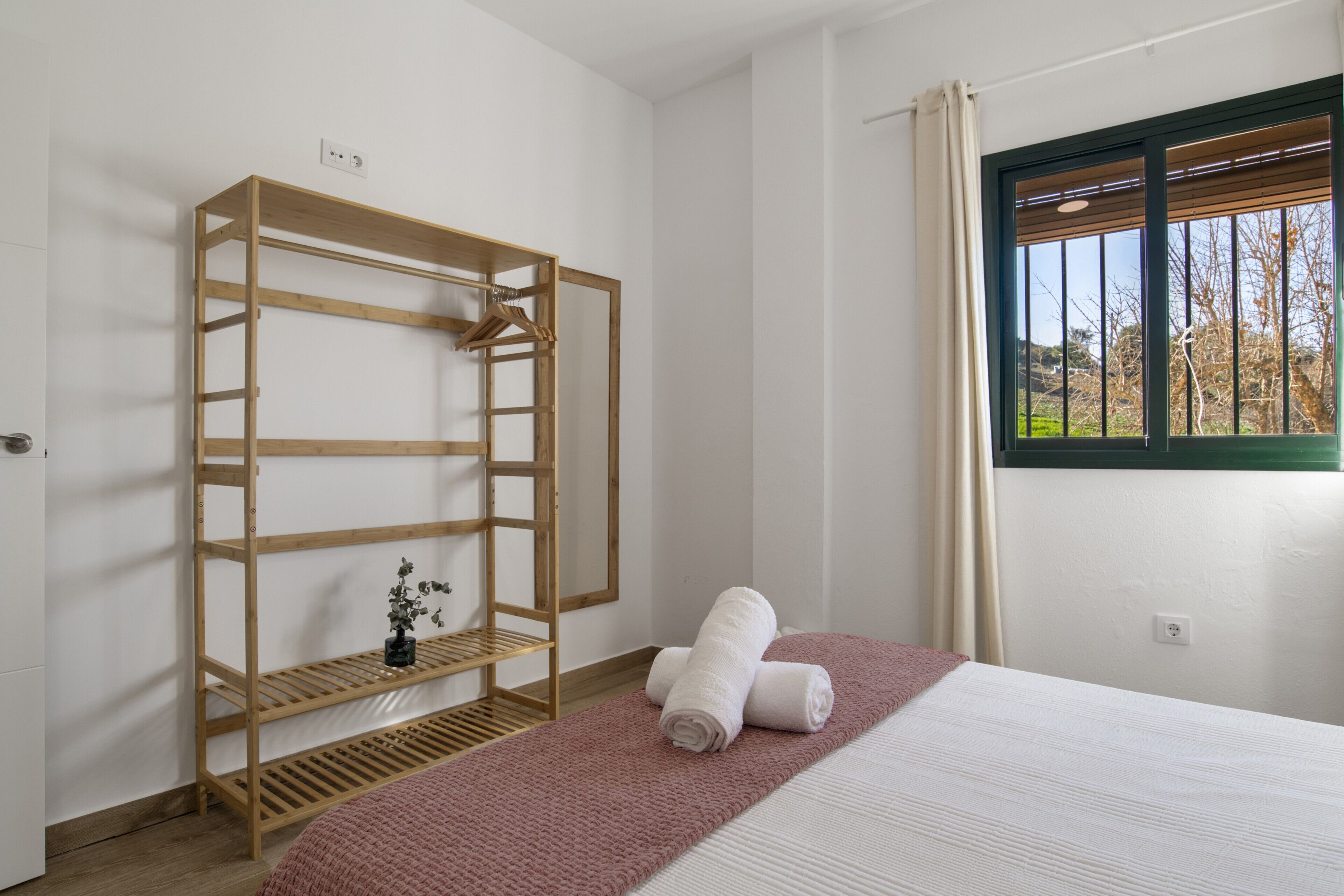 Ein Schlafzimmer mit einem Holzschrank, einem Bett mit weißer Matratze, einer rosa Decke und einem Fenster mit Blick auf Bäume.