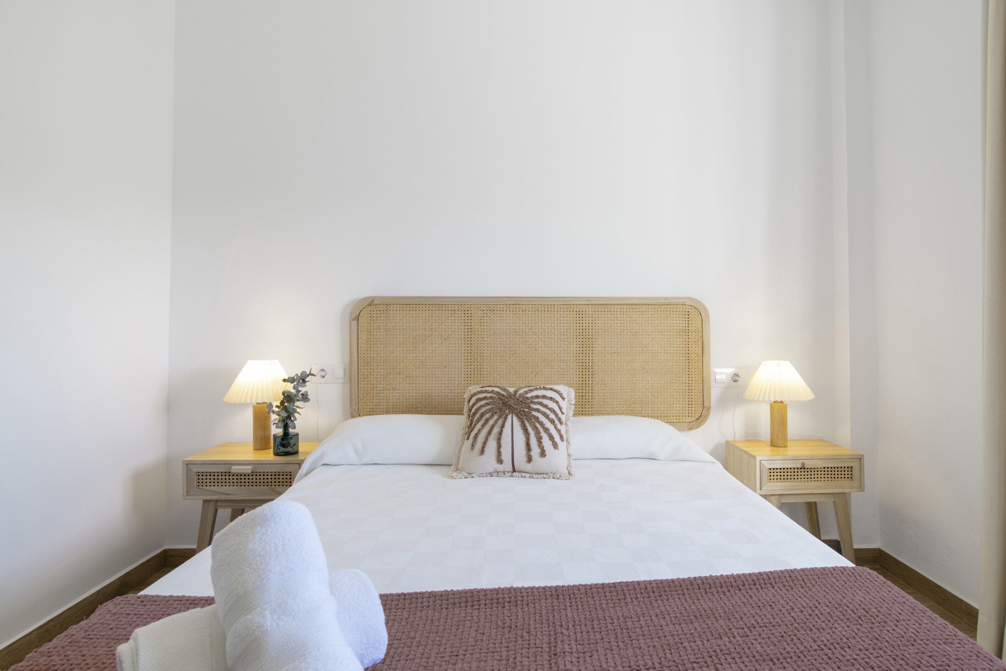 Una cama bien hecha con cabecera, dos mesitas de noche con lámparas y un cojín decorativo.