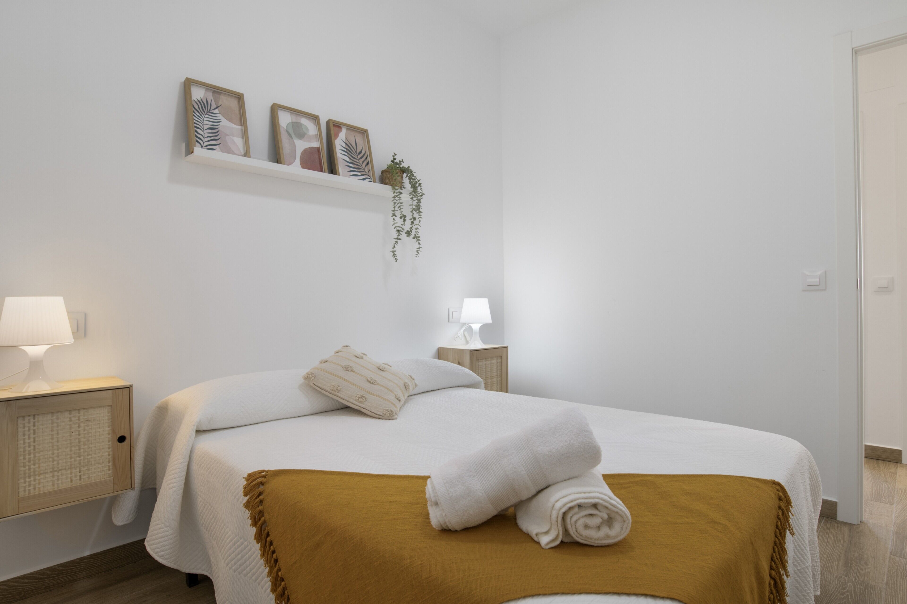 Une chambre à coucher bien rangée, avec un lit, des lampes de chevet, une étagère ornée d’œuvres d’art encadrées et une plante suspendue.