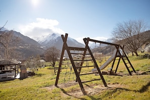 Un parque infantil con columpios y una estructura para escalar en un área verde, con montañas al fondo.