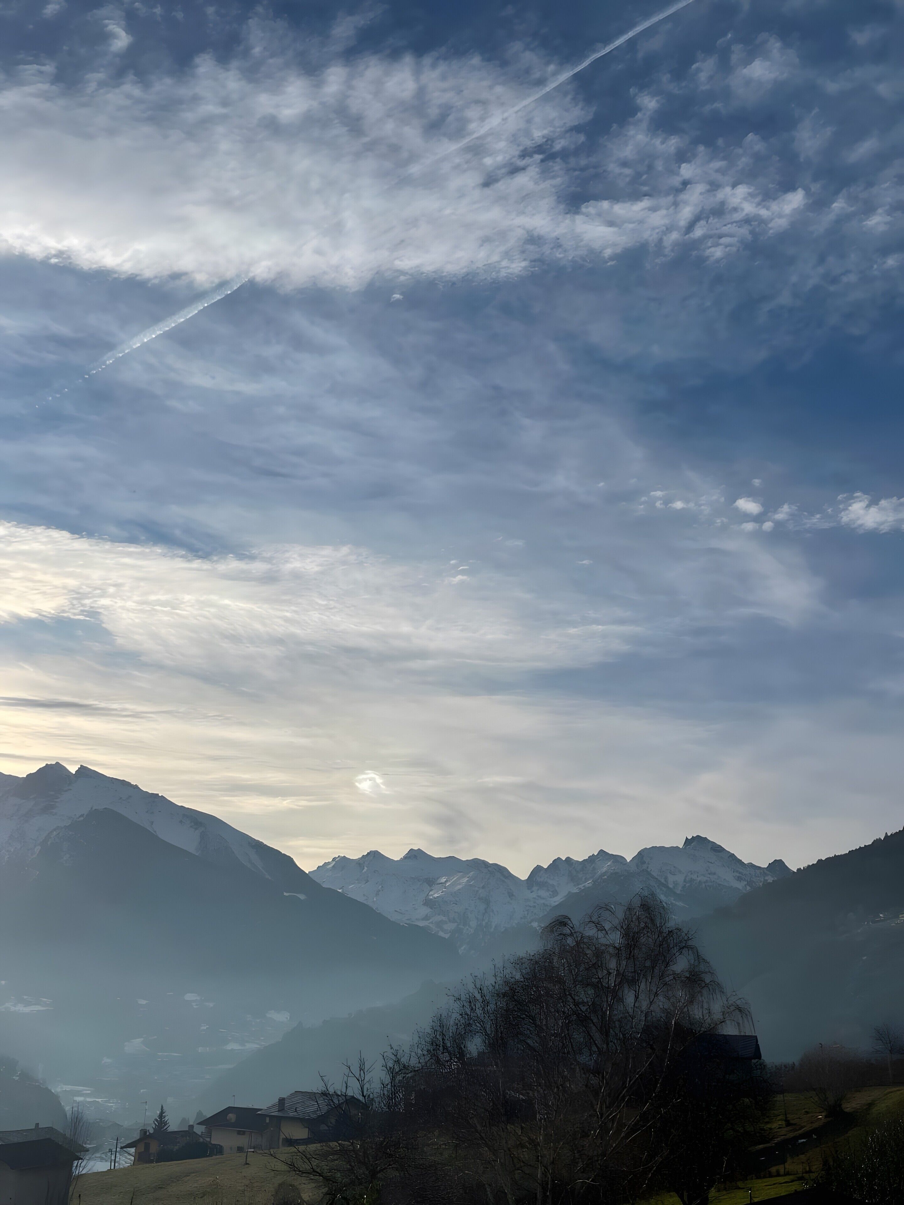 A mountainous landscape with snow-capped peaks, a clear sky, and a visible contrail.