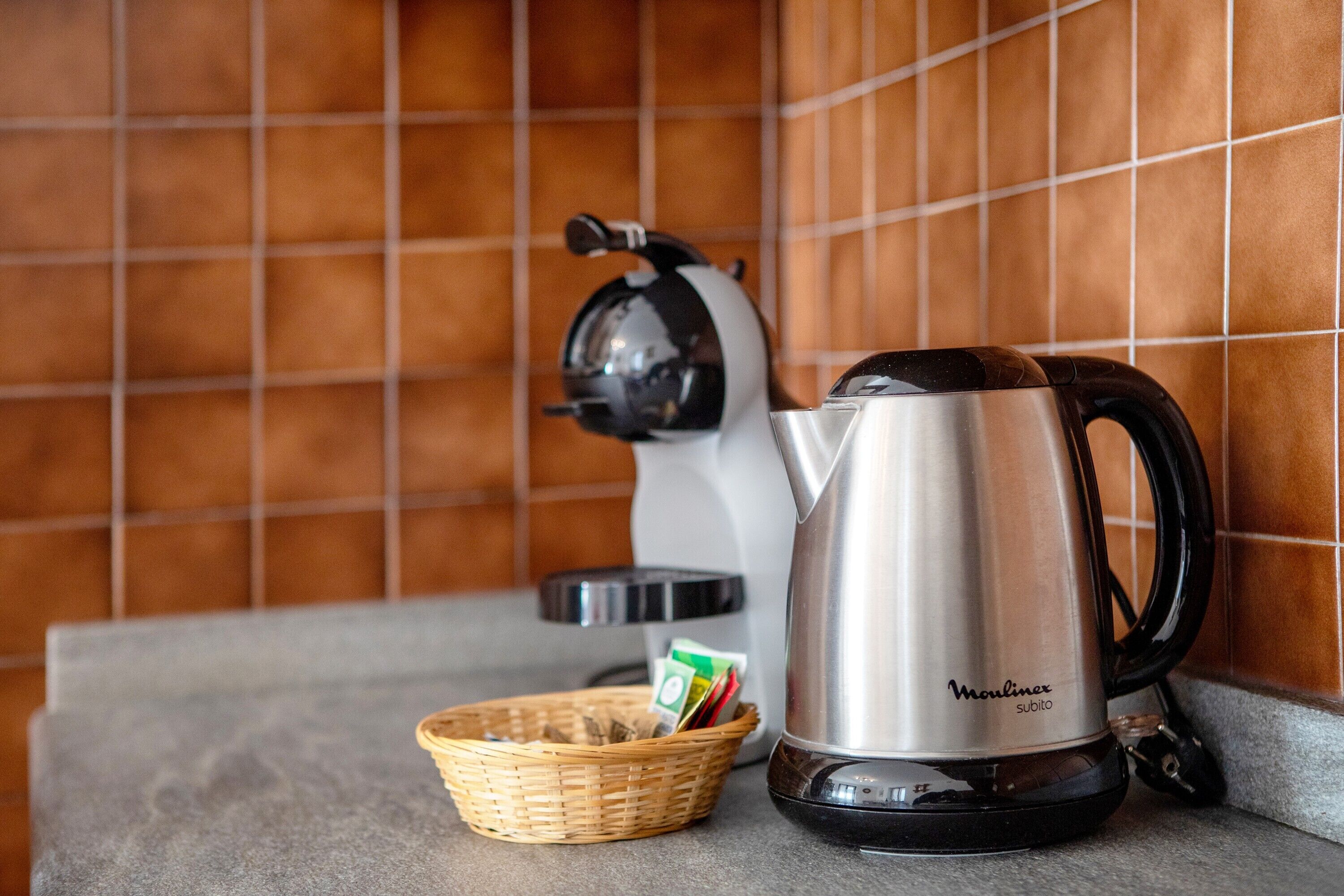 A kitchen with a stainless steel kettle, a coffee maker, and a basket with items on a countertop.