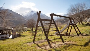 Une aire de jeux avec des balançoires et une structure d’escalade, située dans un espace verdoyant avec des montagnes en arrière-plan.