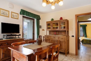 Een eethoek met een houten tafel en stoelen, een televisie op een standaard, een kast met glazen deuren en een raam met gordijnen.