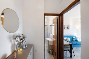 Un interno moderno con uno specchio, un vaso con fiori secchi, una piccola decorazione a forma di casa, una zona pranzo con tavolo e sedie, e un divano blu con un quadro colorato appeso al muro.