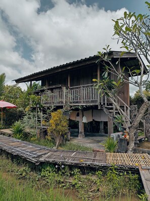 A wooden house with a balcony, surrounded by lush greenery and a small pond.
