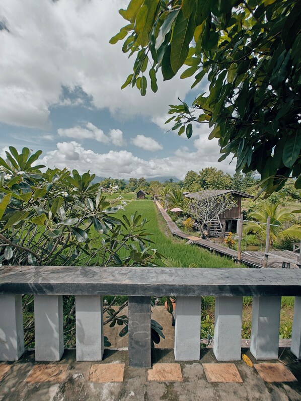 A view from a balcony with a stone railing, overlooking a rural landscape with green fields, a wooden structure, and a clear sky. - hueankwankhao  (Chom Thong)