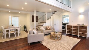 A spacious living area with a sofa, coffee table, and dining set. A staircase leads to an upper level. - 6 Dooley Street Bargara (Bargara)