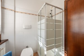 Un bagno con pareti piastrellate di bianco, cabina doccia in vetro, un water bianco e una porta in legno.