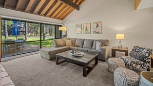 A spacious living room with a large sectional sofa, a coffee table, and a patterned armchair. There are two side tables with lamps, and a glass door leading to an outdoor area with a table and chairs. - Cozy Condo Near the VIllage (Sunriver)