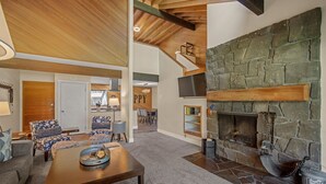 A spacious living area with a stone fireplace, wooden ceiling beams, and a cozy seating arrangement. - Cozy Condo Near the VIllage (Sunriver)