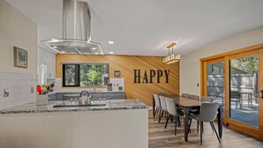 A modern kitchen with a central island, a dining area with a wooden table and chairs, and a large window with a view of greenery. - Cozy Condo Near the VIllage (Sunriver)