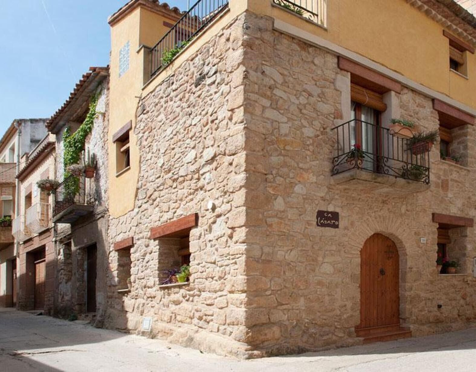 Un edificio de piedra con puerta de madera, un balcón con barandilla de metal y una ventana pequeña con plantas.