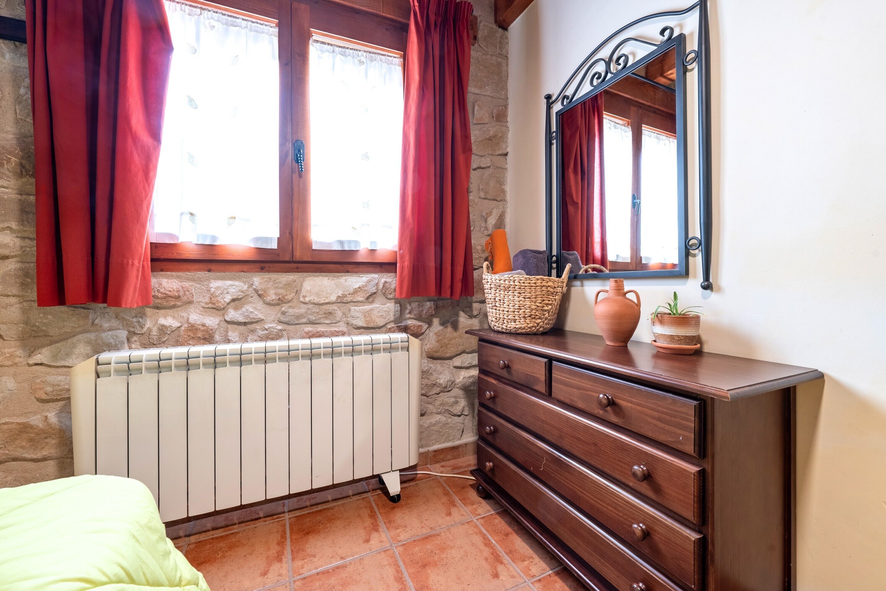 Una habitación con pared de piedra, cómoda de madera, espejo, radiador y ventana con cortinas rojas.