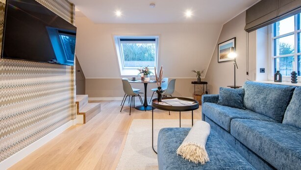 A modern living room with a blue sofa, a dining table with chairs, a staircase, and a large window providing natural light. - F27 Living Whg. 08 - F27 Living (Sylt)