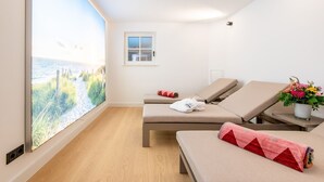 A room with two reclining chairs, a window with a beach view, a small table with a potted plant, and a window with a view of a building. - F27 Living Whg. 08 - F27 Living (Sylt)