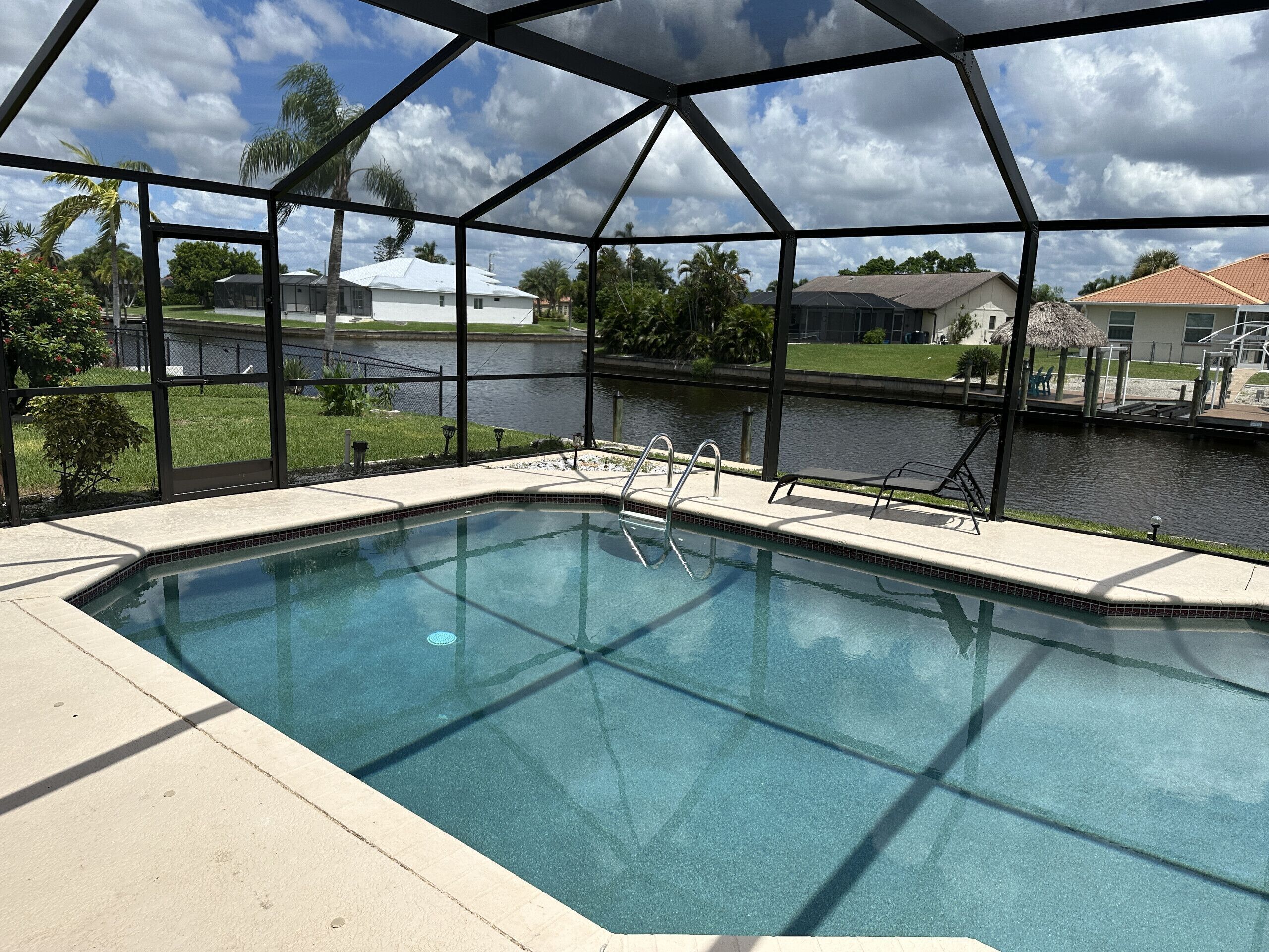A swimming pool with a metal roof structure, surrounded by a fence, with a view of a body of water and houses in the background.